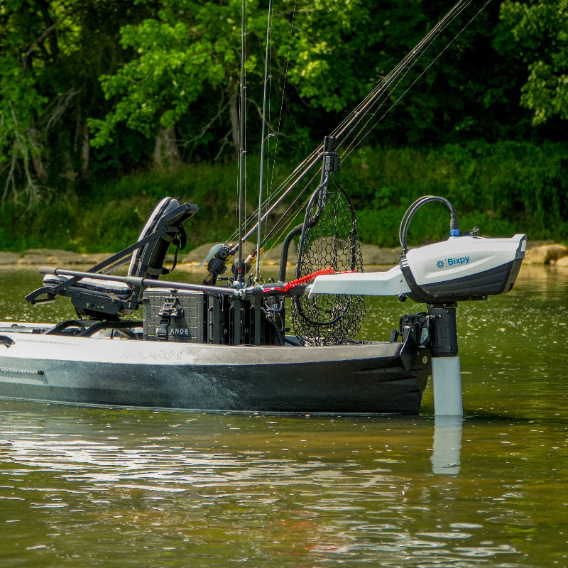 Grey and white fishing kayak with a Bixpy electric outboard motor, fishing rods, and net, floating in murky water with green trees along the shoreline.