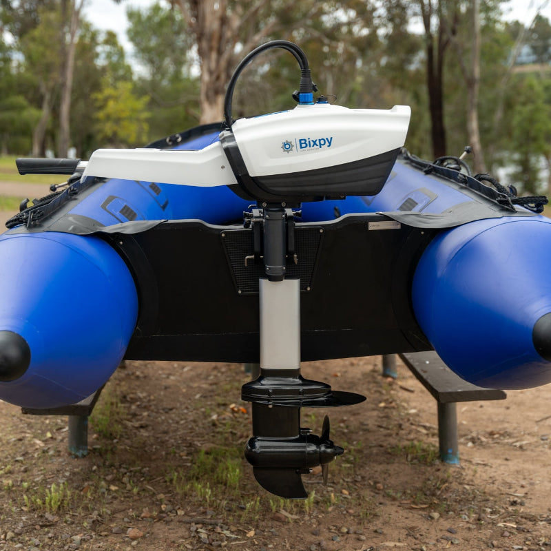 White Bixpy electric outboard motor with silver shaft and propeller mounted on the stern of a blue and black inflatable boat on land.