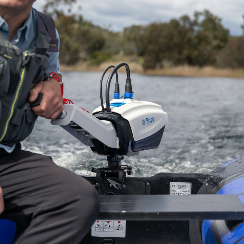 Close-up of a white Bixpy electric outboard motor mounted on an inflatable boat, showing cables connected and a person's hand on the tiller handle as it propels through water.