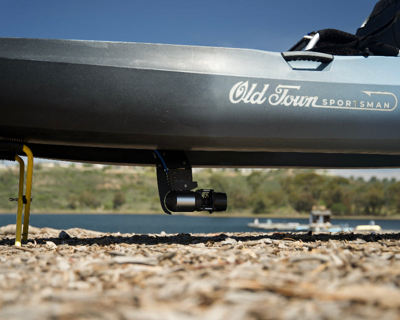 Bixpy Jet electric motor mounted to the underside of a dark grey Old Town Sportsman kayak resting on a stand on a rocky shore next to water.