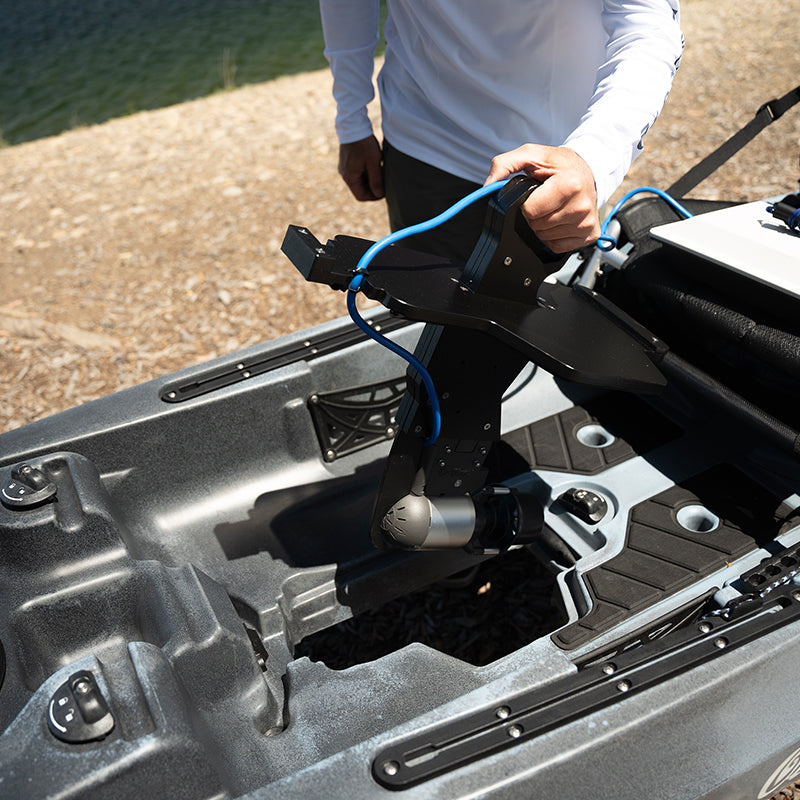 Top-down view of a person installing a Bixpy Jet electric motor with a black adapter into the pedal well of a grey kayak on a rocky shore.