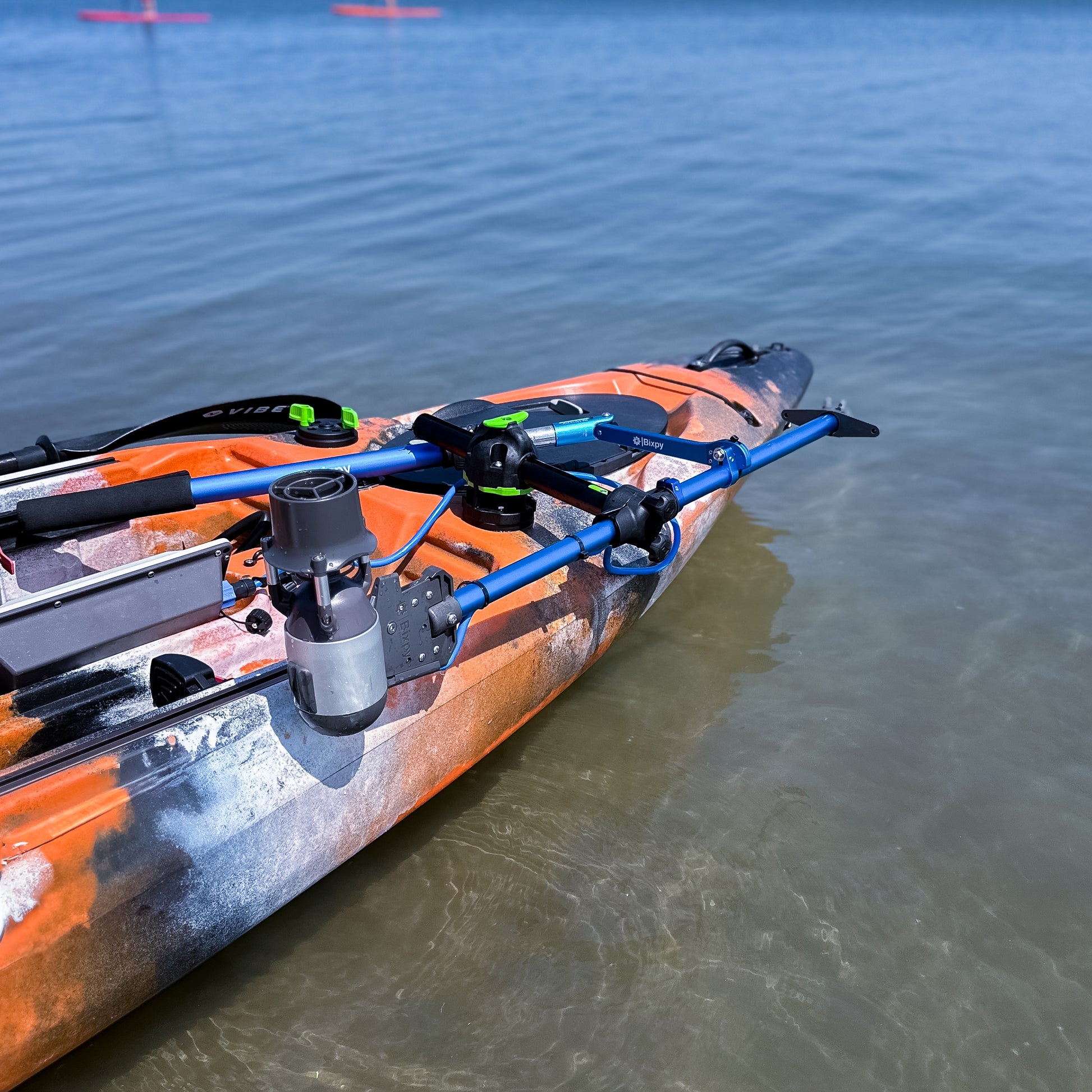 Bixpy Jet electric motor mounted on a blue pole mount adapter at the front of an orange camouflage kayak, submerged in shallow clear water.