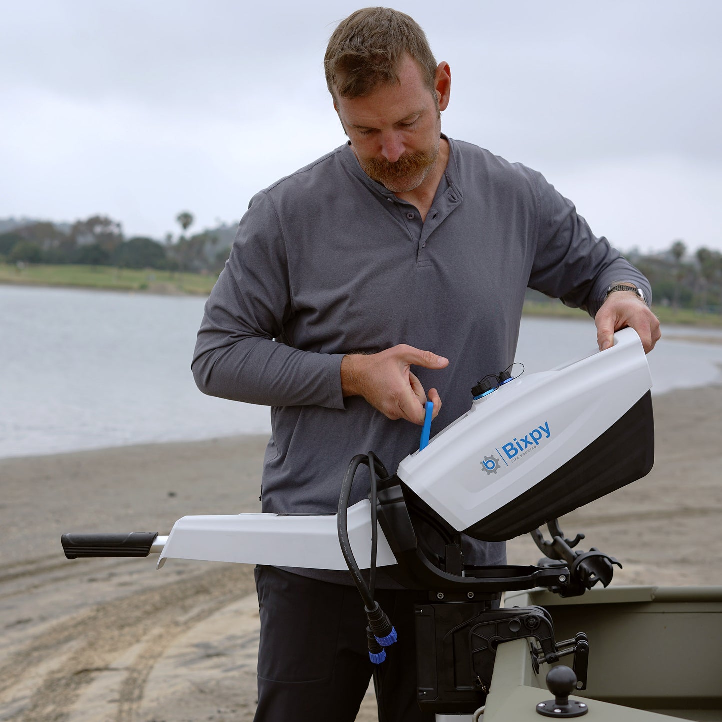 Man on a sandy beach adjusting a white Bixpy electric outboard motor mounted to a small olive green boat.