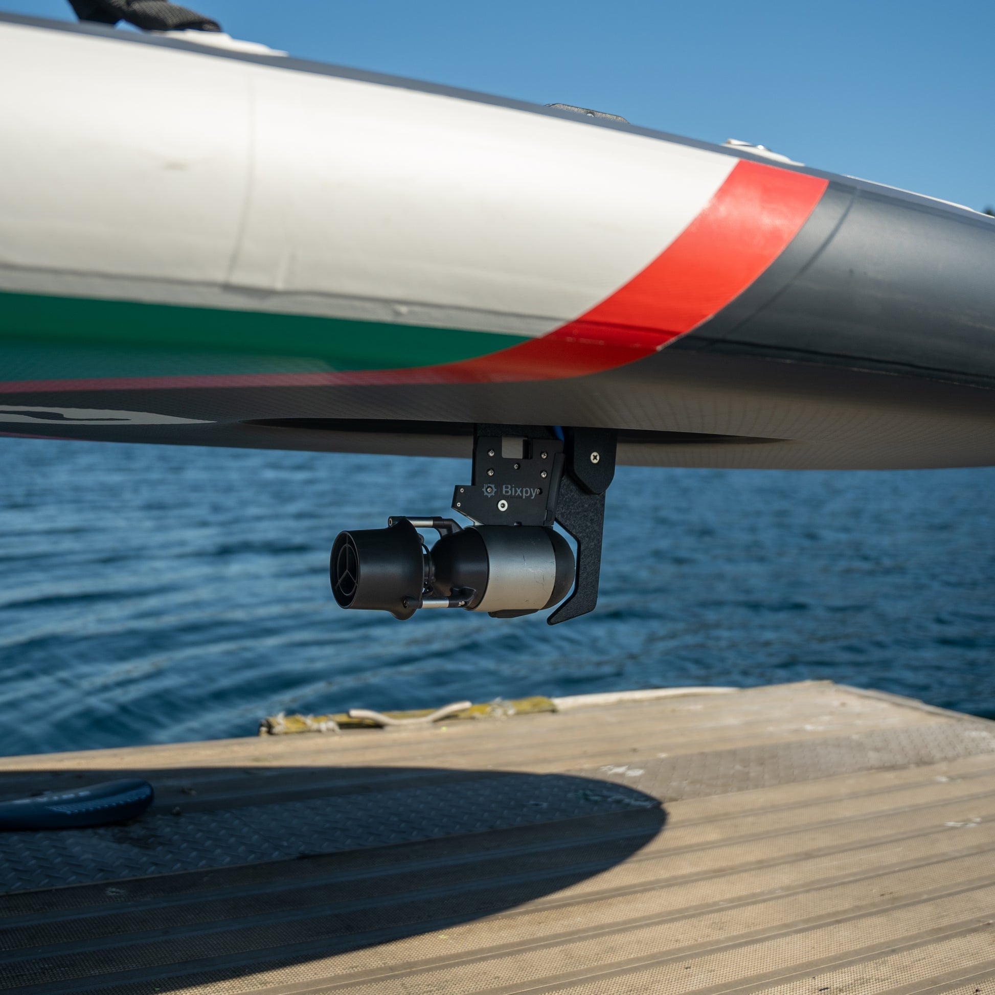 Close-up of a Bixpy Jet electric motor mounted beneath a white and black boat with a red stripe, positioned above calm blue water.