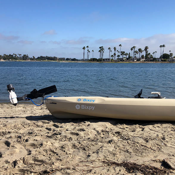 Light tan kayak with a white Bixpy Jet electric motor mounted on the stern and a blue cable, beached on a sandy shore by calm water and palm trees.