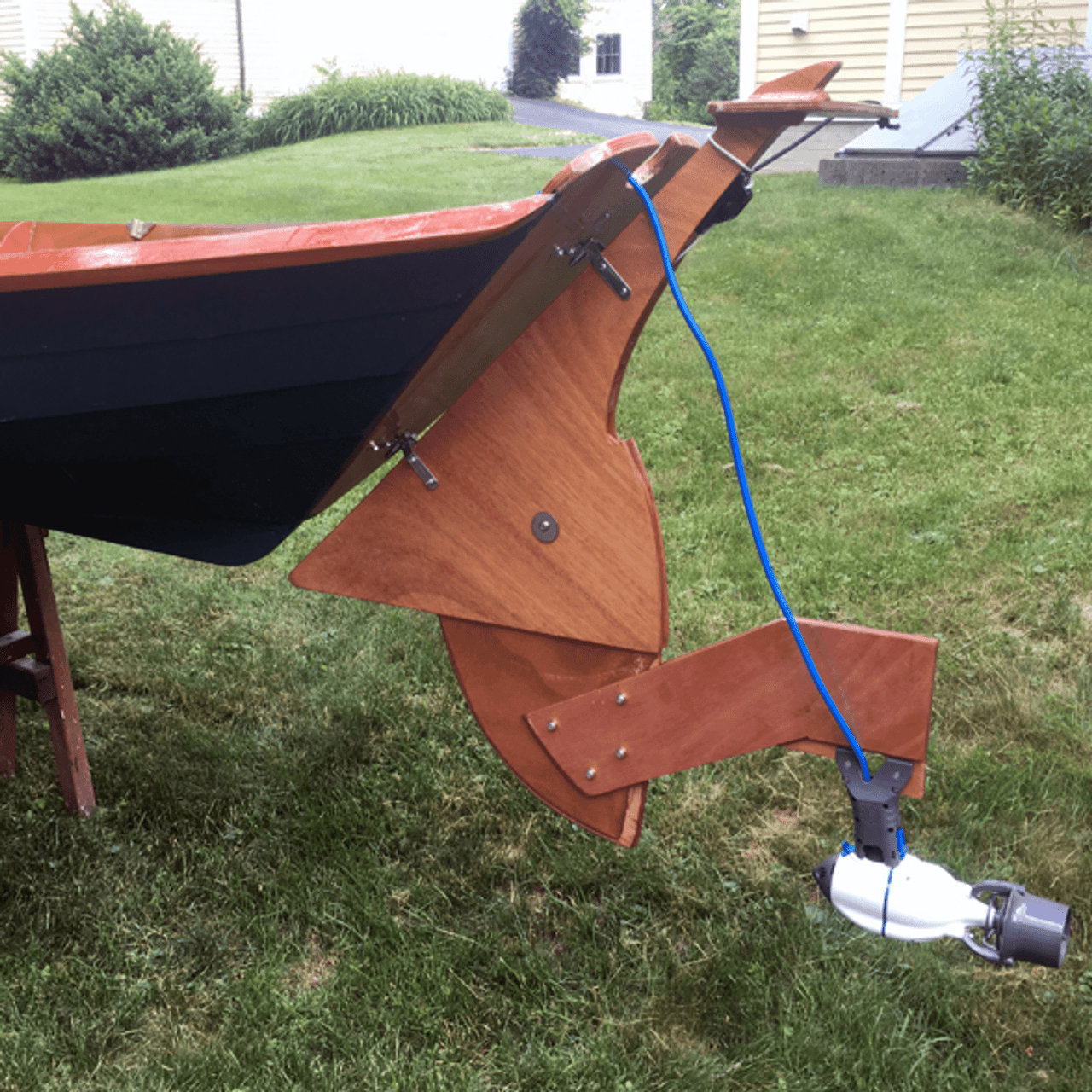 Wooden boat fitted with a custom wooden rudder system, featuring a Bixpy motor mounted at the bottom with a blue power cable attached, displayed on grass in a residential yard.