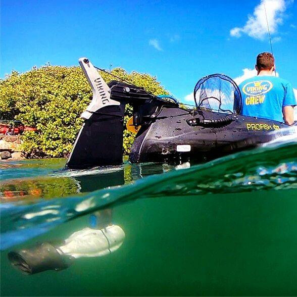 Side view of a Viking Profish kayak equipped with a Bixpy motor and rudder system in the water, showing both above and below the surface, with a person wearing a “Viking Crew” shirt standing on board.