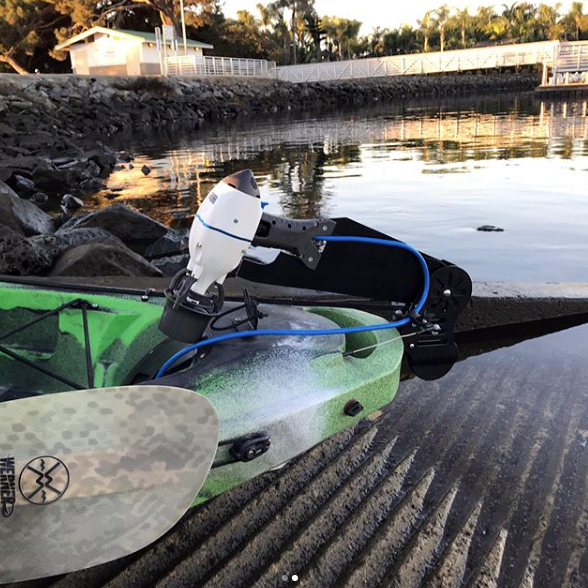 Green kayak equipped with a Bixpy motor mounted on a rudder steering adapter, positioned at a boat ramp near calm water with a shoreline and trees in the background.