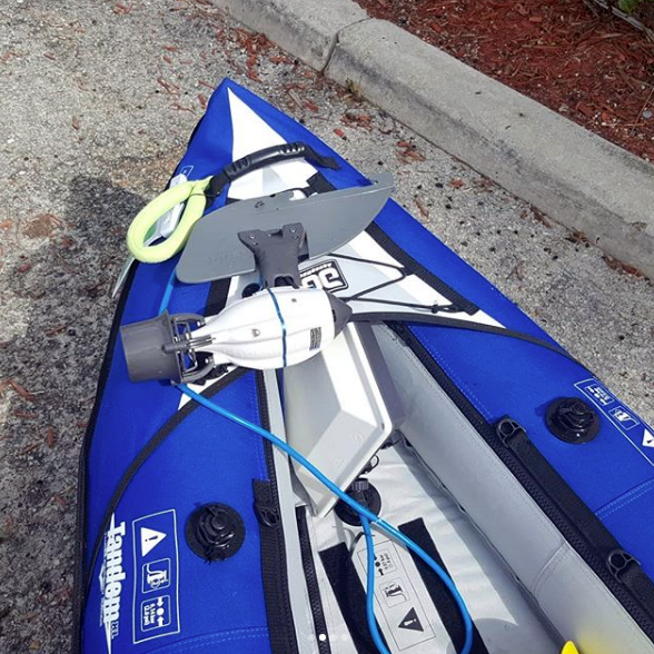 Top-down view of a blue inflatable kayak fitted with a Bixpy motor and fin adapter, with a blue power cable and paddle secured on the deck, parked on a concrete surface.