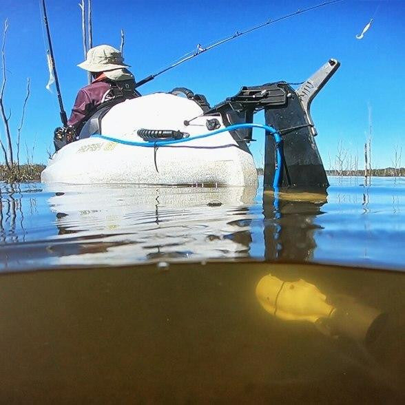 Angler in a white fishing kayak on calm water, equipped with a Bixpy motor mounted on a rudder system; the photo captures both above and below the surface, showing the submerged motor.