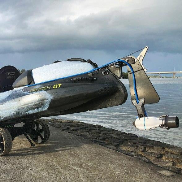Black Viking Profish GT kayak on a wheeled cart, equipped with a Bixpy motor mounted on a large rudder, positioned on a concrete ramp beside a calm waterway under cloudy skies.