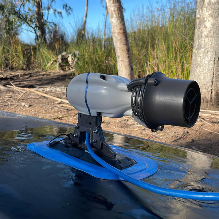 White Bixpy Jet electric motor mounted on a black Glue-On Fin Box Adapter with a blue adhesive patch, on a dark inflatable watercraft in an outdoor natural setting.