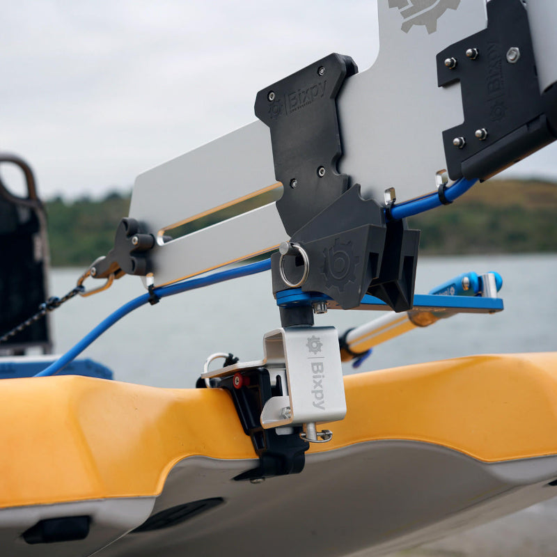 Close-up of a Bixpy rudder mounting adapter system with silver blade, black clamps, and blue components, installed on the stern of an orange kayak on water.