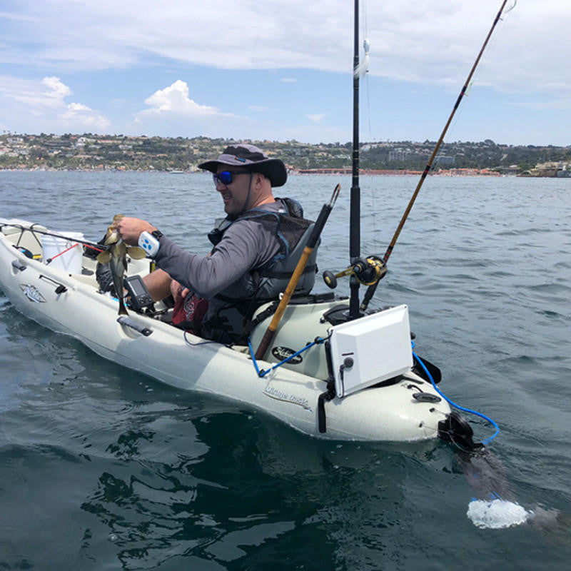 Angler in a beige Mirage Outback kayak, holding a fish, propelled by a Bixpy electric motor with battery box, on choppy water near a distant coastline.