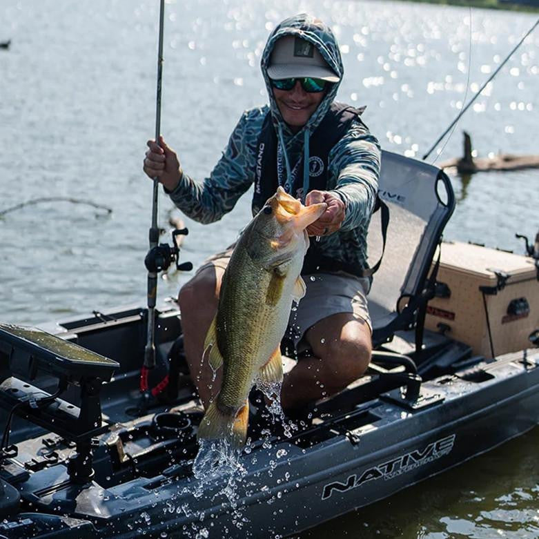 Angler in a grey Native Watercraft kayak holding up a large fish that is splashing water, with a fishing rod nearby.