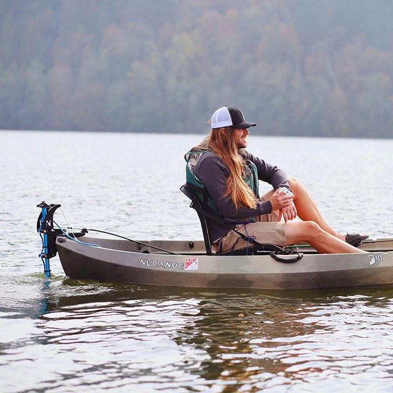 Man in a grey Nucanoe kayak propelled by a Bixpy Jet electric motor mounted on a blue vertical pole, on calm water with a forested shoreline.
