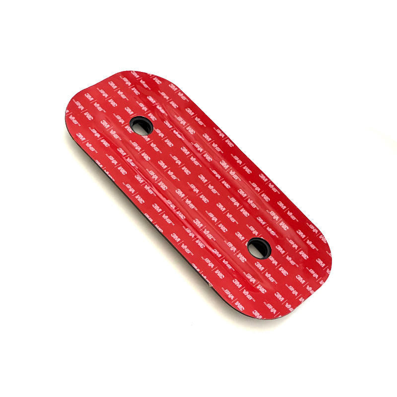 Underside view of a black oval adhesive mounting pad with red 3M adhesive backing and two circular holes on a white background.