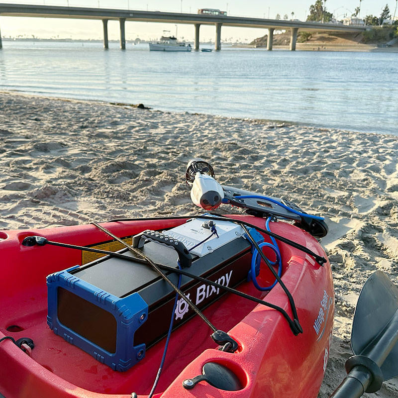Bixpy Jet Motor and Battery Pack installed on a red kayak on a sandy beach. A bridge and boat are visible in the background.