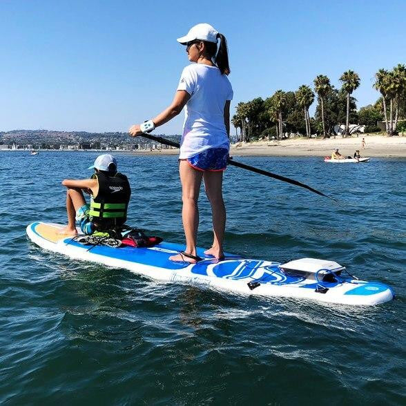 A woman paddleboarding with a child on a Bixpy-powered SUP near a beach on a sunny day.