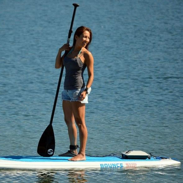 Woman smiling on a Bixpy-powered stand-up paddleboard, holding a paddle on calm water.