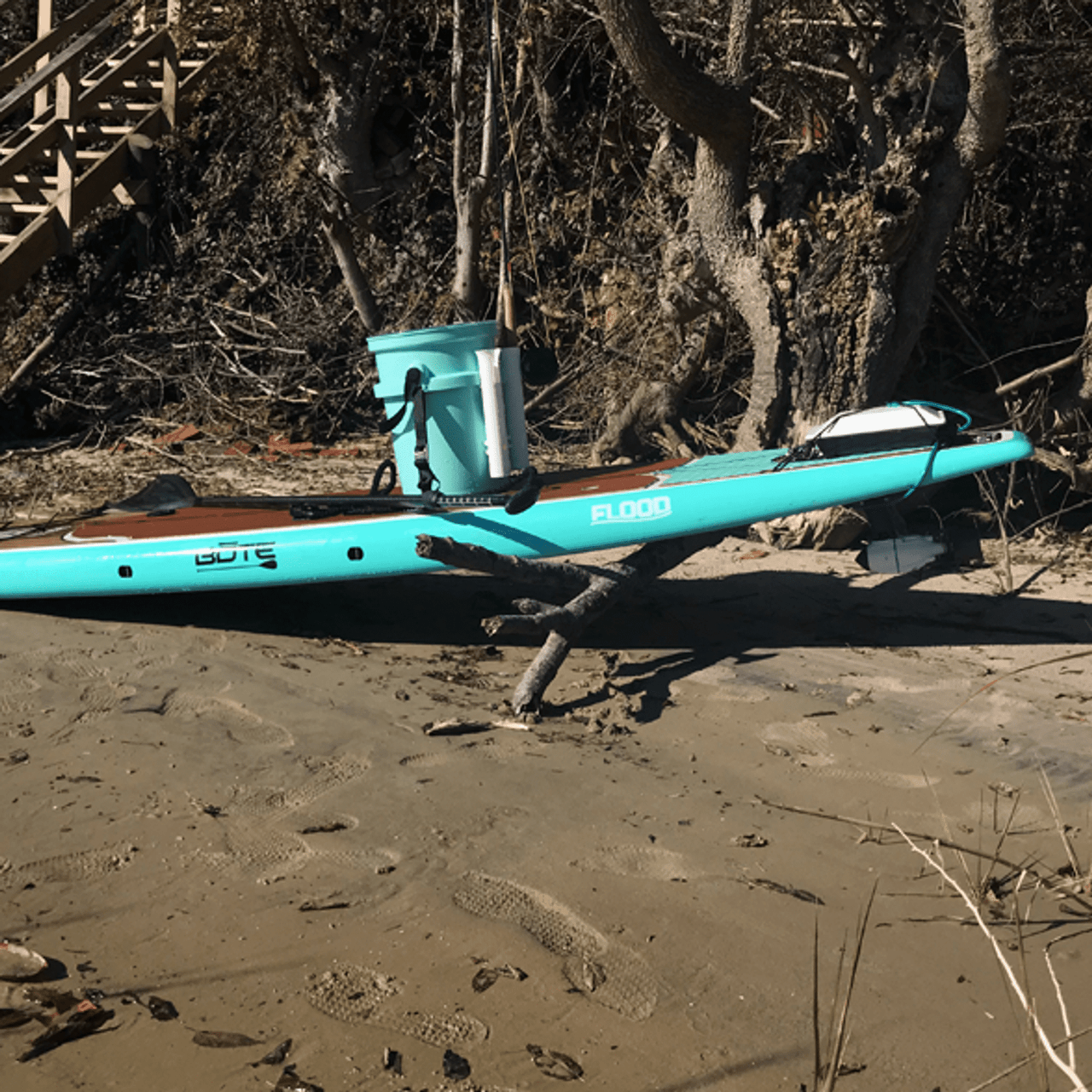 Turquoise BOTE Flood paddleboard resting on a sandy shoreline, equipped with a Bixpy motor mounted at the rear and a gear bucket secured to the deck, positioned near driftwood and trees.
