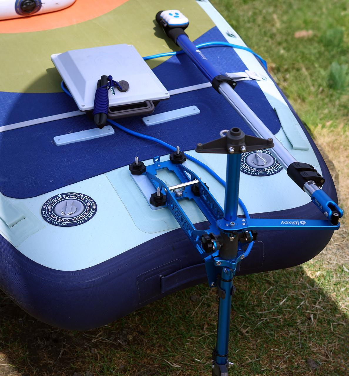 Blue and green inflatable watercraft with a mounted Bixpy pole mount adapter, a Bixpy battery, and a paddle, resting on grass.