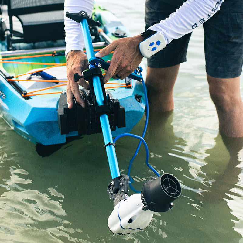 Person in shallow water adjusting a white Bixpy Jet electric motor on a blue and black universal transom mount attached to a blue kayak, with a remote control on their wrist.