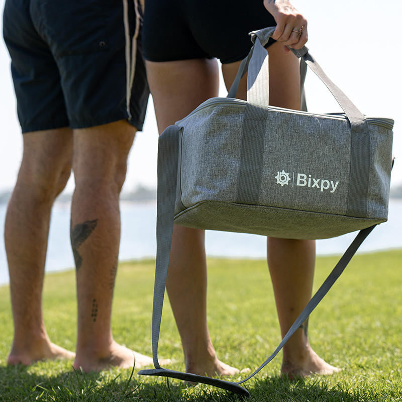 Two people holding the Bixpy Travel Bag by the handles, walking barefoot on grass.
