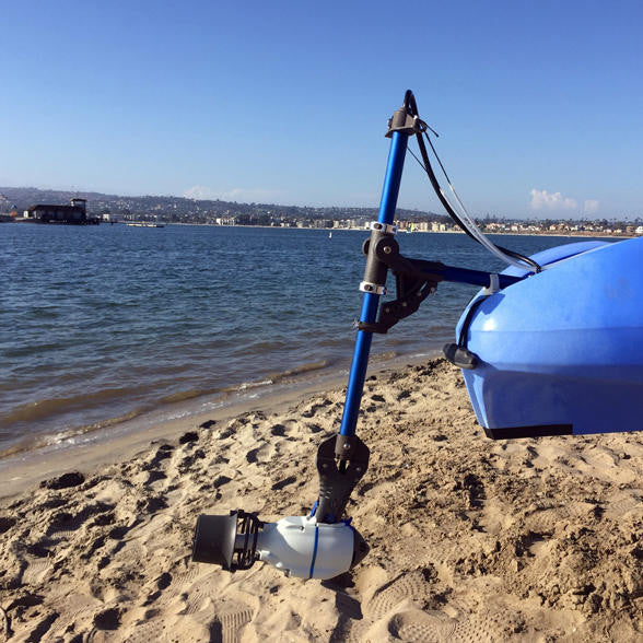 Blue kayak on a sandy beach equipped with a Bixpy Universal Kayak Adapter for J-1 Motors, showing the motor mounted and ready for launch with water and distant shoreline in the background.