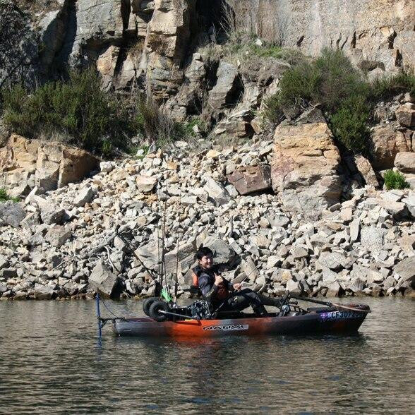 Angler fishing from an orange and black kayak equipped with a Bixpy Universal Kayak Adapter and motor, positioned on calm water near a rocky cliff shoreline.