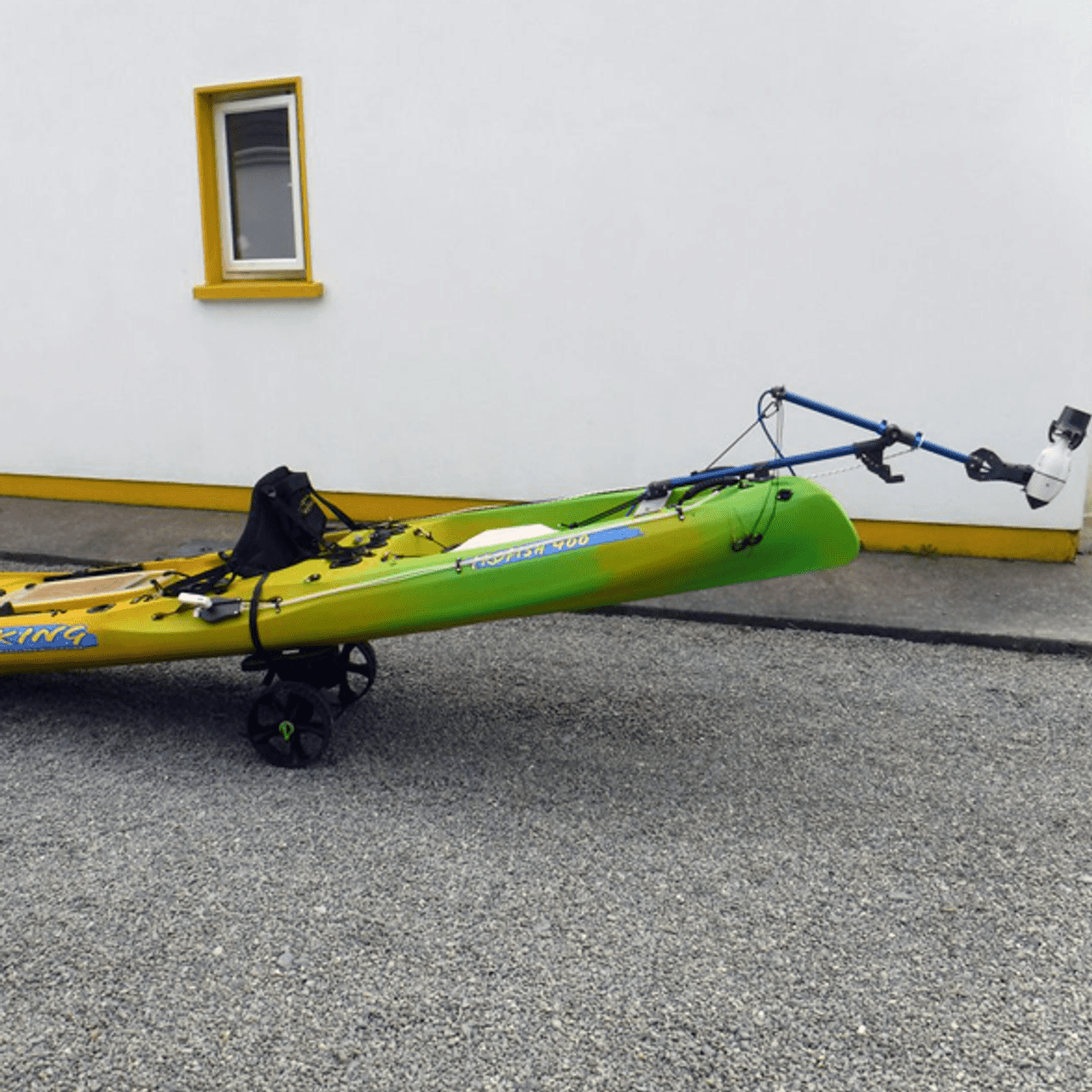 Viking Profish 400 kayak on a wheeled cart, outfitted with a Bixpy Universal Kayak Adapter and motor, parked on gravel in front of a white and yellow building.