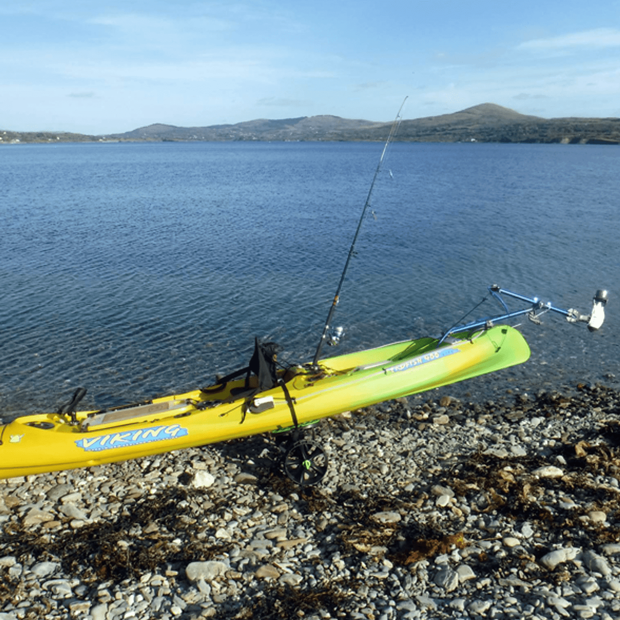 Yellow and green Viking Profish 400 kayak equipped with a Bixpy Universal Kayak Adapter and motor, beached on a rocky shoreline with fishing rods mounted and calm blue water in the background.