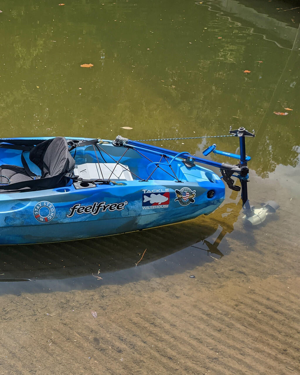 Blue camouflage Feelfree kayak with Bixpy Universal Kayak Adapter and motor, operating in very shallow murky water near a sandy bank.