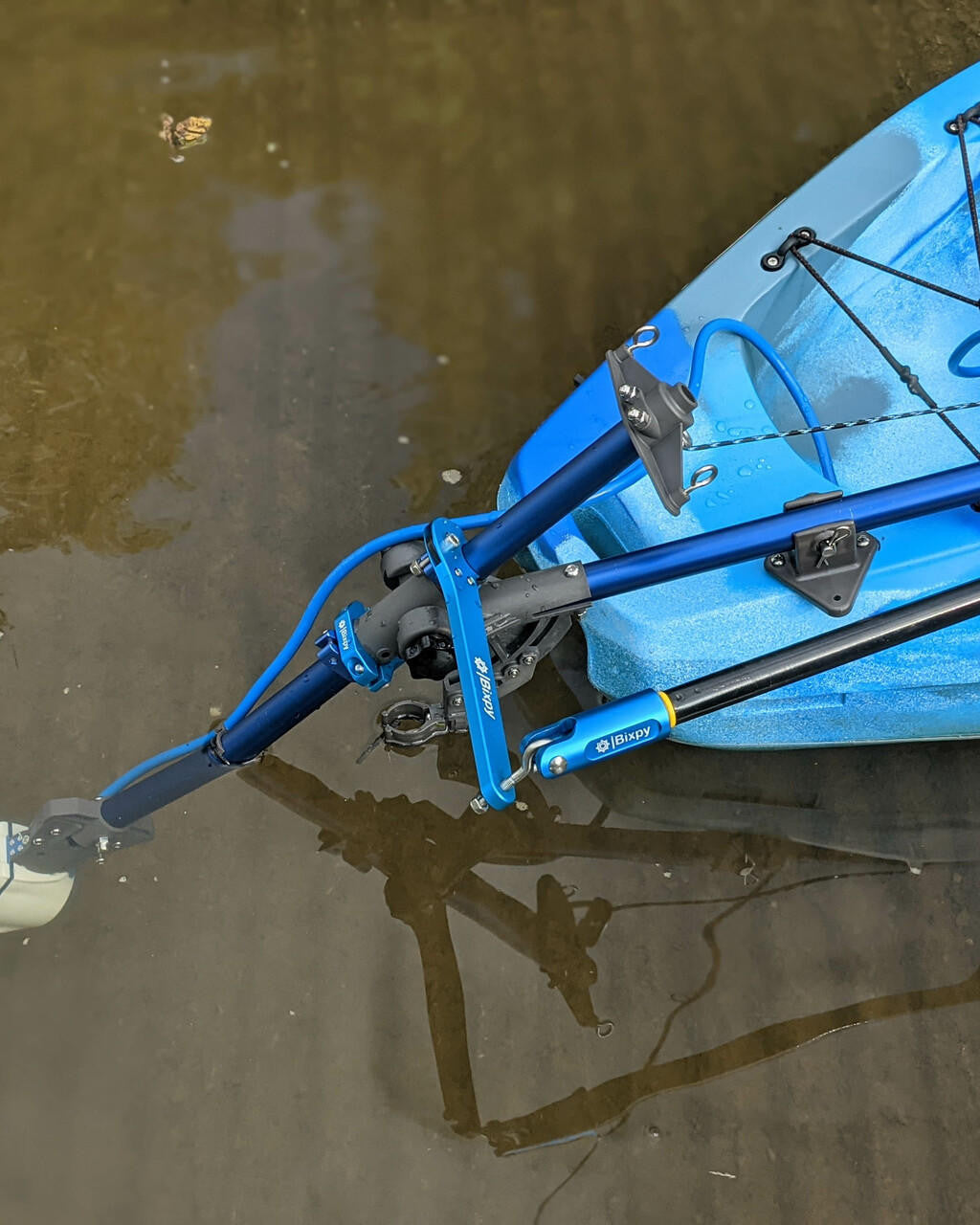 Close-up of a Bixpy Jet electric motor mounted to a blue kayak via a blue Universal Kayak Adapter, partially submerged in water with reflections.