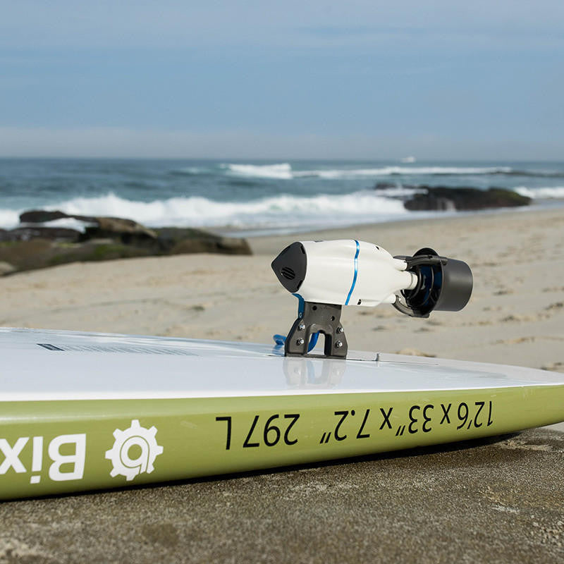 White Bixpy Jet electric motor with blue cable mounted on the front deck of a green and white paddleboard, on a sandy beach.