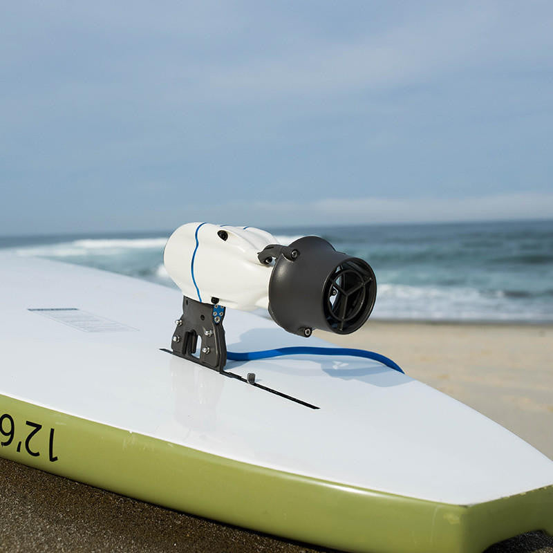 White Bixpy Jet electric motor with blue stripe mounted on a green and white Bixpy branded paddleboard, on a sandy beach with ocean waves.