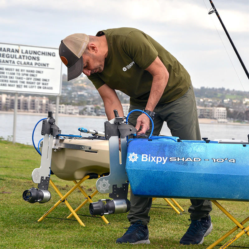 Man in Bixpy shirt preparing a tan kayak and a blue SHAD 10'4" kayak, both equipped with Bixpy Jet electric motors and mounts, on yellow stands near water.