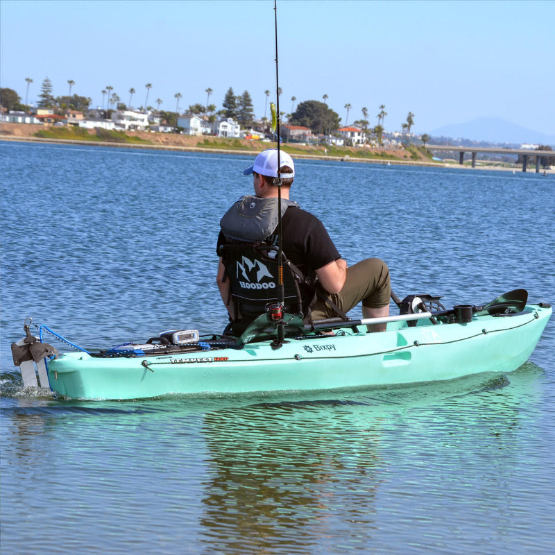 Person in a kayak propelled by a Bixpy Jet electric motor, fishing on calm water with houses and a bridge in the distance.