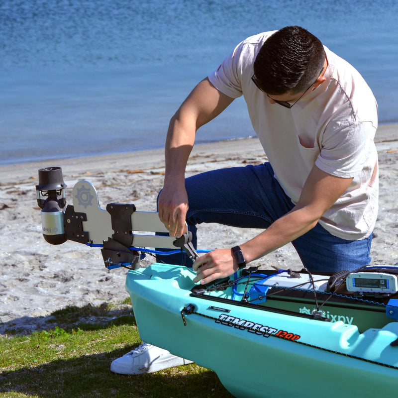 Man kneeling on grass adjusting a Bixpy Jet electric motor mounted on a light gray rudder adapter at the stern of a blue Tempest 12'0 kayak on a sandy beach.