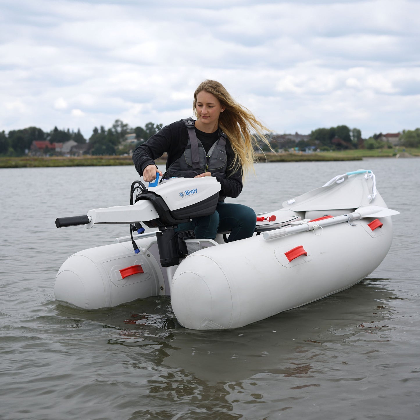 Woman in a white inflatable pontoon boat adjusting a Bixpy electric outboard motor in rippled water with houses in the distance.