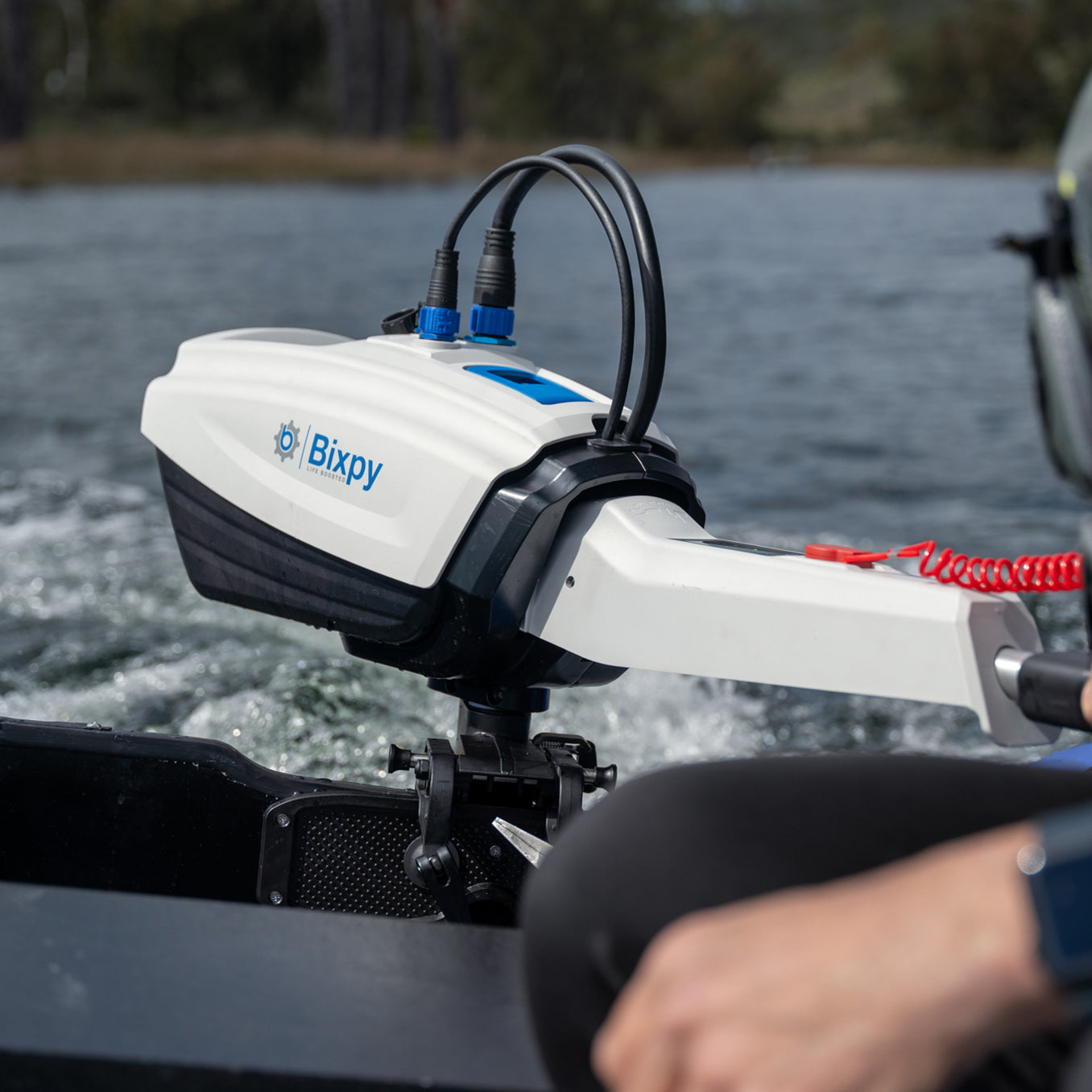 Close-up of a white and black Bixpy electric motor battery pack mounted vertically on a black bracket attached to a blue inflatable tube, with cables connected and water in background.