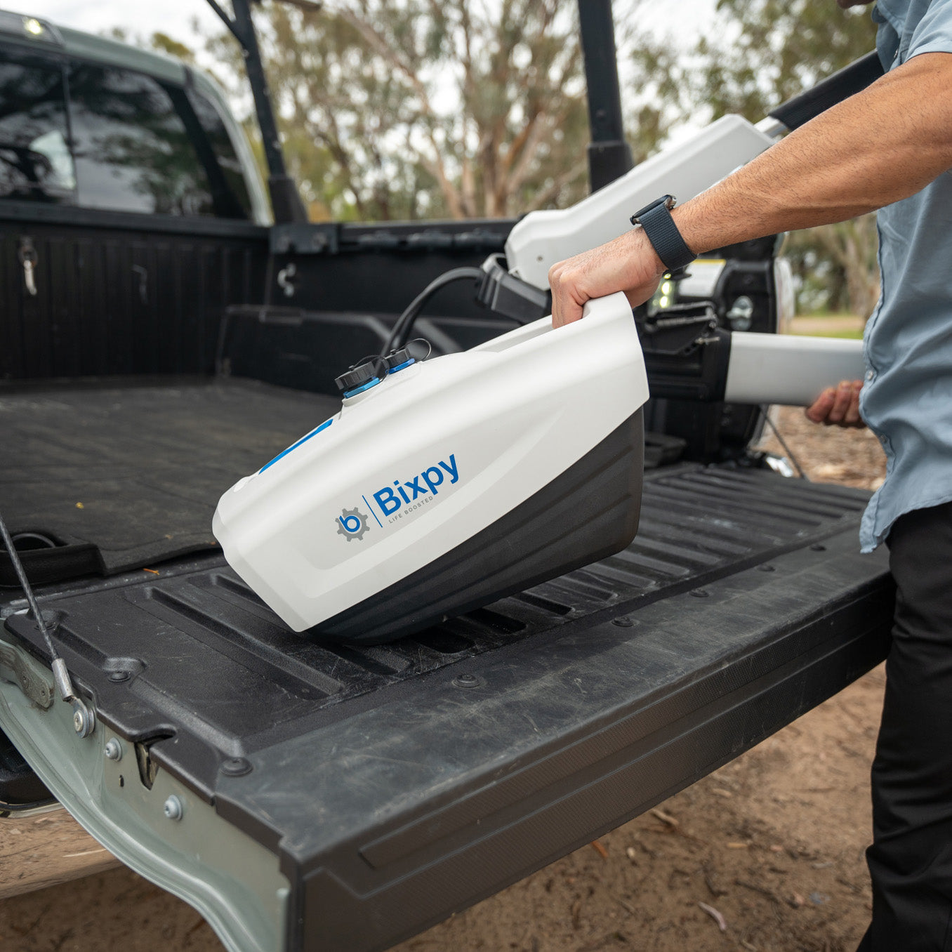 Person placing a white and black Bixpy electric motor battery pack into the bed of a pickup truck, with other Bixpy equipment visible.