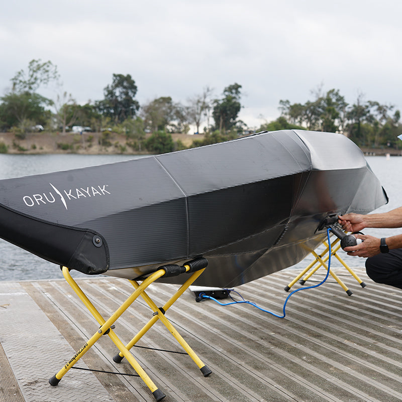 Person's hands attaching a Bixpy motor to a dark grey Oru folding kayak resting on yellow stands on a wooden dock.
