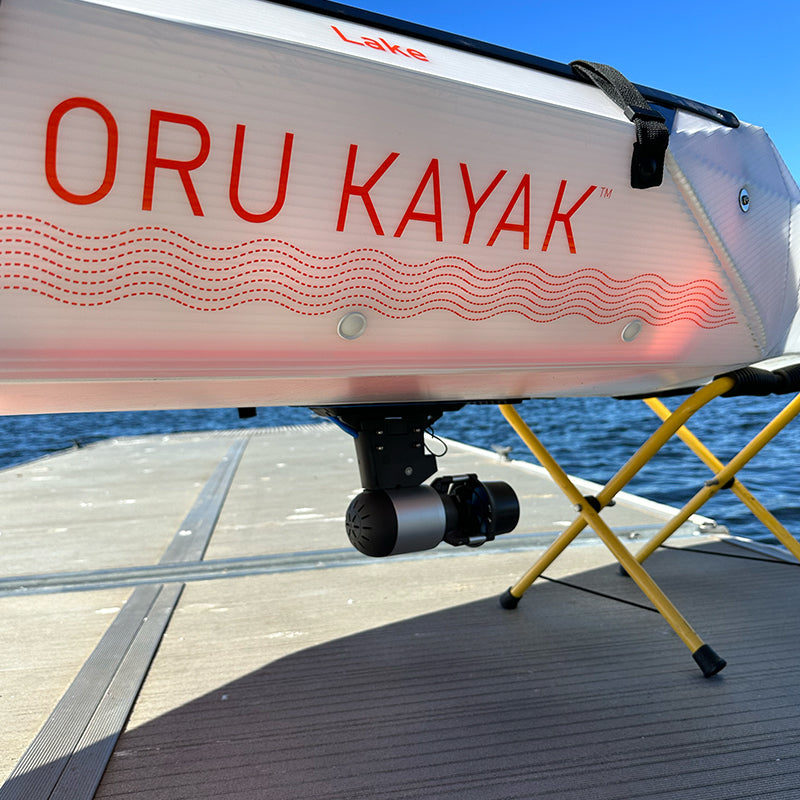 White Oru folding kayak with a Bixpy Jet electric motor mounted underneath, resting on yellow stands on a wooden dock with calm water in the background.