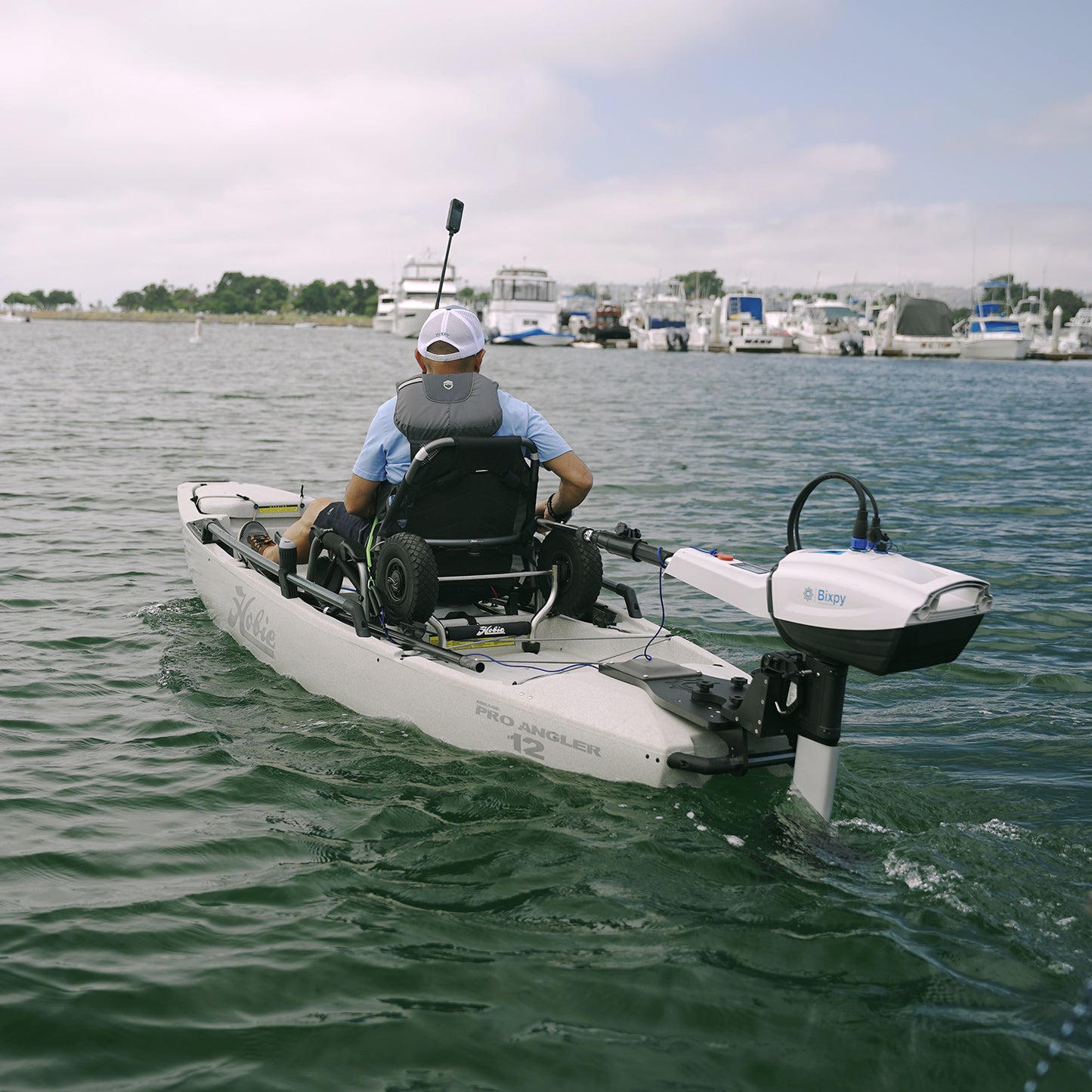 A person in a Hobie Pro Angler 12 kayak equipped with a wheelchair seat paddles away from the camera on calm water. Mounted on the back of the kayak is a Bixpy N-3 outboard motor, installed using the Bixpy Power Pole to Transom Conversion Adapter. In the background, boats and yachts are docked at a marina under a partly cloudy sky.
