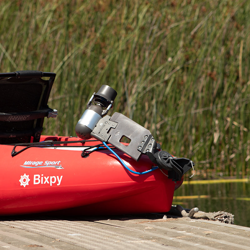 Red Mirage Sport kayak with a Bixpy Jet electric motor mounted using a dark grey specialized adapter, resting on a wooden dock with reeds in the background.