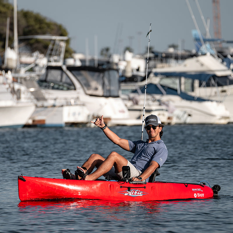 Man in a red Hobie kayak with a Bixpy electric motor giving a shaka sign, propelling across calm marina waters with boats in the background.