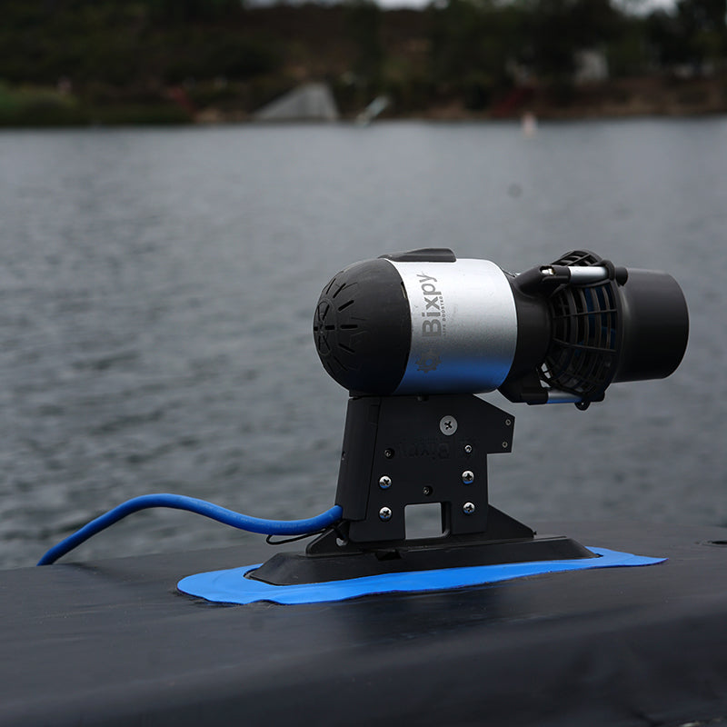Bixpy Jet electric motor mounted on a black Glue-On Fin Box Adapter, secured with a blue adhesive patch on a dark inflatable watercraft, with blurred water in the background.