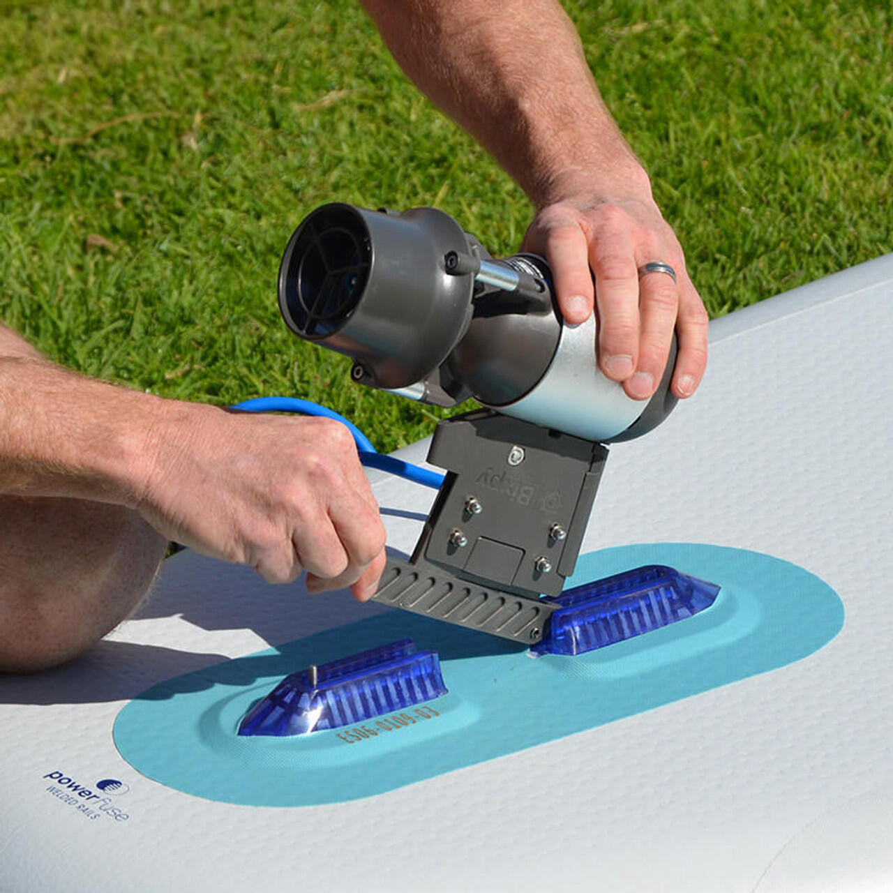 Close-up of a person's hands installing a Bixpy Jet electric motor with a grey adapter into two blue fin boxes on a white paddleboard on grass.