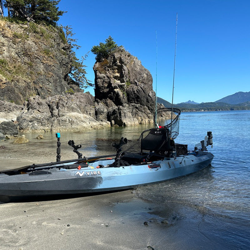 Blue and gray Vibe fishing kayak with a Bixpy electric motor and multiple fishing rods, beached on a sandy shore next to clear water with rocky cliffs and mountains.